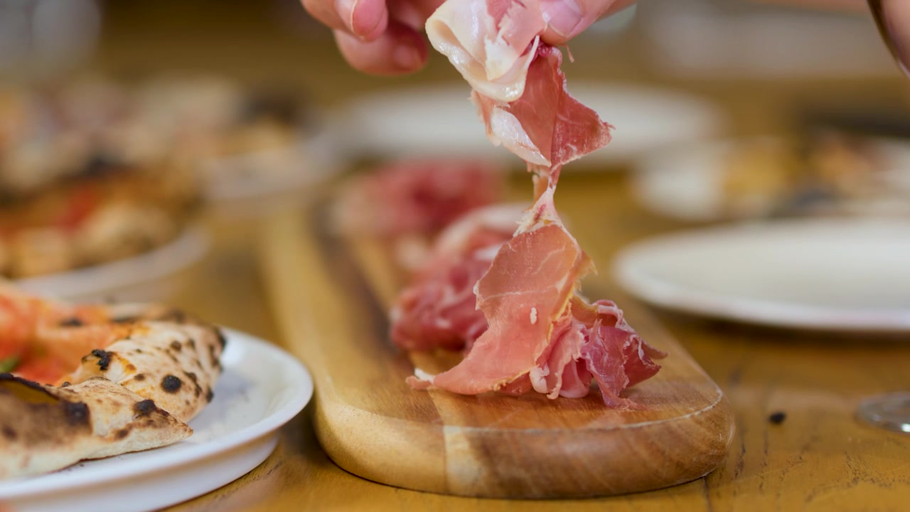 Primo piano di una mano che solleva salumi da un tagliere di legno durante una cena italiana.