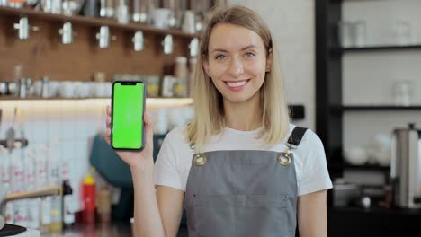 close up portrait of a young woman barista standing in front of the counter in bar and showing green screen of smartphone to camera holding on right hand in cafe.