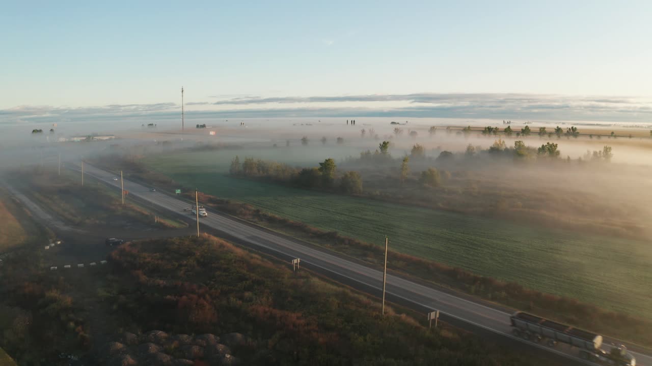 Soft golden morning light shines on fog hanging low over highway and surrounding land. Early morning mist on farm fields with trees poking out above. Aerial view from drone with traffic on road.