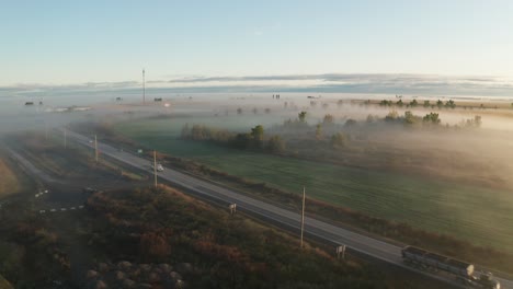 soft golden morning light shines on fog hanging low over highway and surrounding land. early morning mist on farm fields with trees poking out above. aerial view from drone with traffic on road.