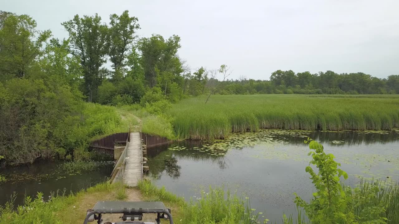 imágenes de drones de 4k de un hombre irreconocible en un utv de lado a lado junto a un pequeño río con nenúfares y cielos nublados, árboles, puente peatonal rural por sendero de camping utv en mid michigan orv trails