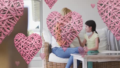 animation of hearts over caucasian woman and her daughter wearing face masks playing with pet dog