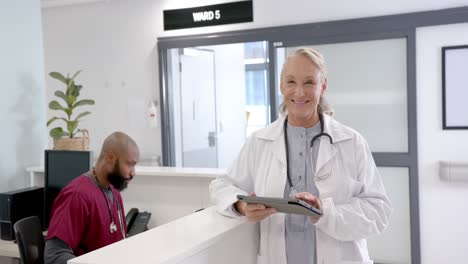 portrait of happy senior caucasian female doctor using tablet in hospital reception, slow motion