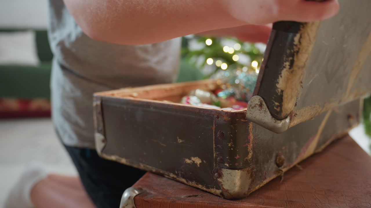 Partial view of boy in grey top and black shorts kneeling while opening old vintage box filled with colorful ornaments beside Christmas tree in bright cozy room preparing for festive celebration