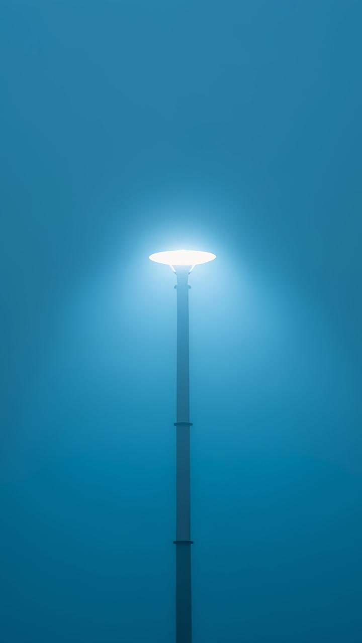 Video verticale: lampione stradale segmentato e luminoso con testa a palo che proietta un alone nella nebbia blu su una strada deserta