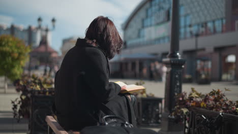 woman seated outdoors in black coat reading book, hair partially covering face, flipping pages, flower pots sway in breeze. urban street features blurred storefronts and lamp pole