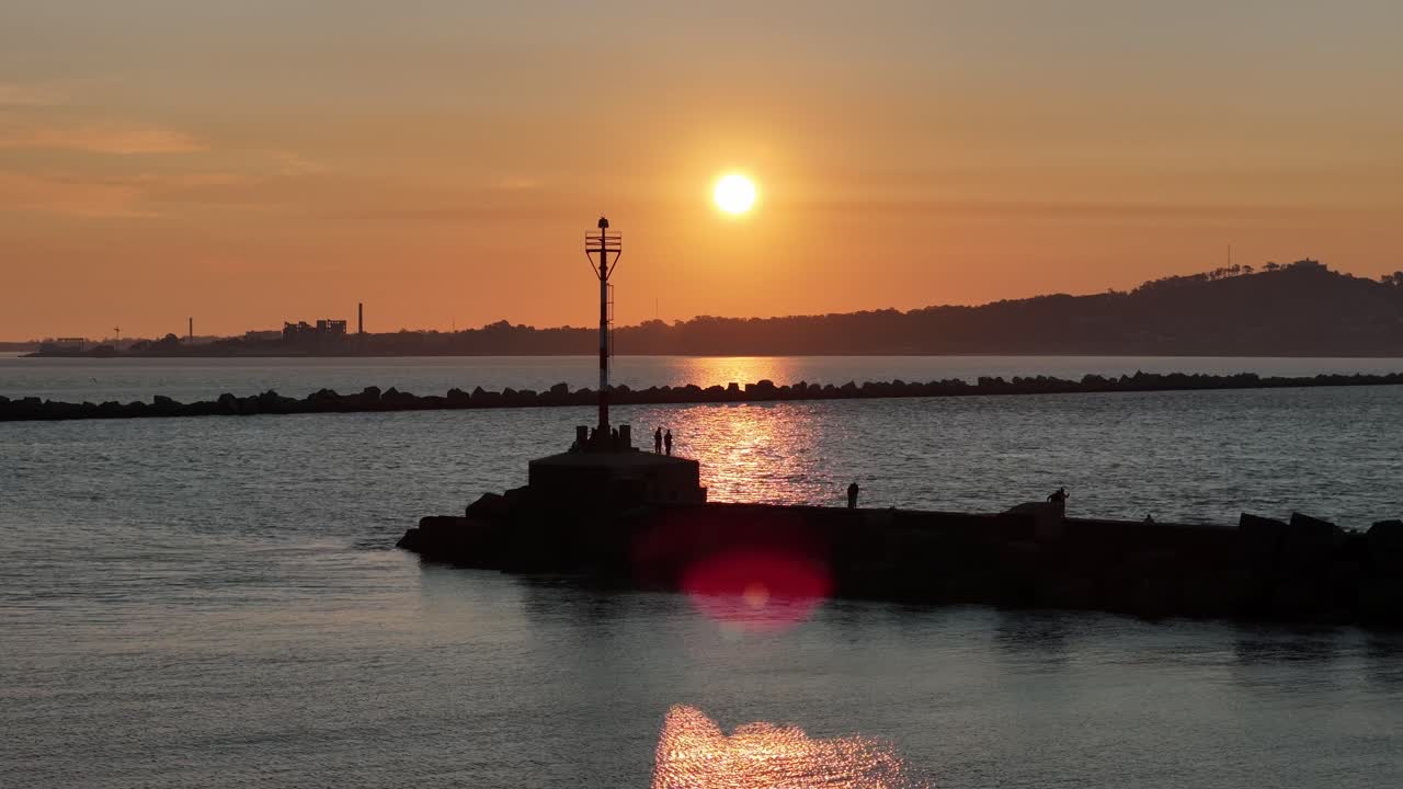 Drone shot at sunset over Montevideo Bay, showing the breakwater with silhouetted fishermen and warm winter light reflecting on the water