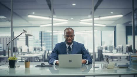 successful black businessman in tailored suit working on laptop computer on top floor office overlooking big city. professional ceo managing environmental, social and corporate governance