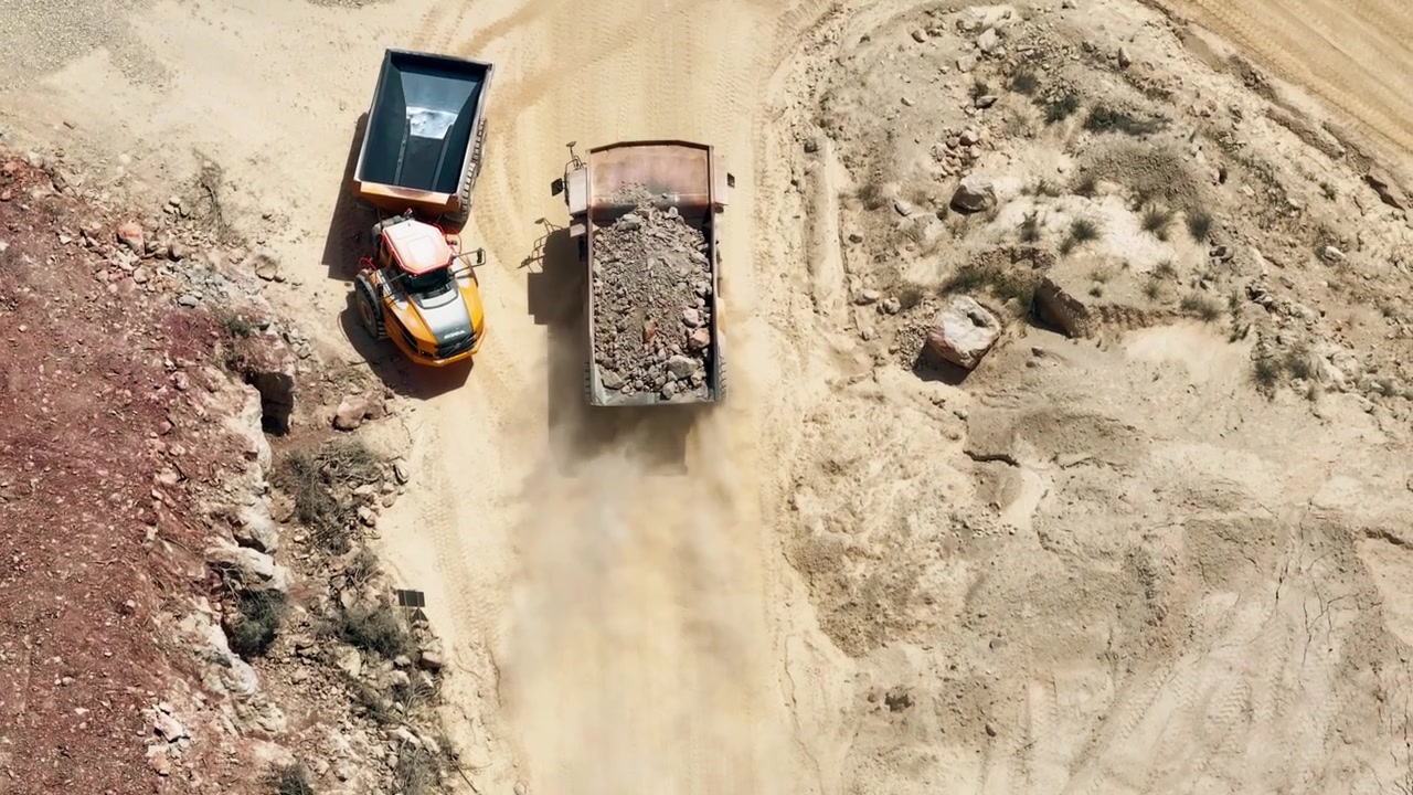 Large dump trucks transport materials in an active limestone quarry. Conveyor belts and heavy equipment process raw materials. Quarry logistics are visible from an aerial perspective.