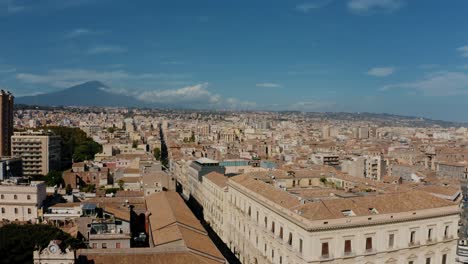flying sideways above the main street in catania with mount etna in the background on a sunny day. aerial city view in sicily with historical buildings.