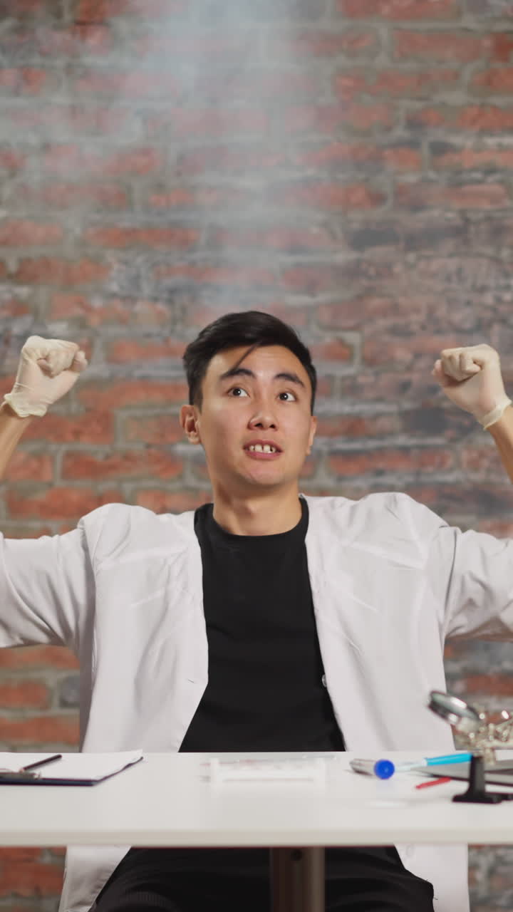 Despaired young Asian scientist in white coat with steam cloud above head crazy of experiment fail sits at workplace in scientific laboratory