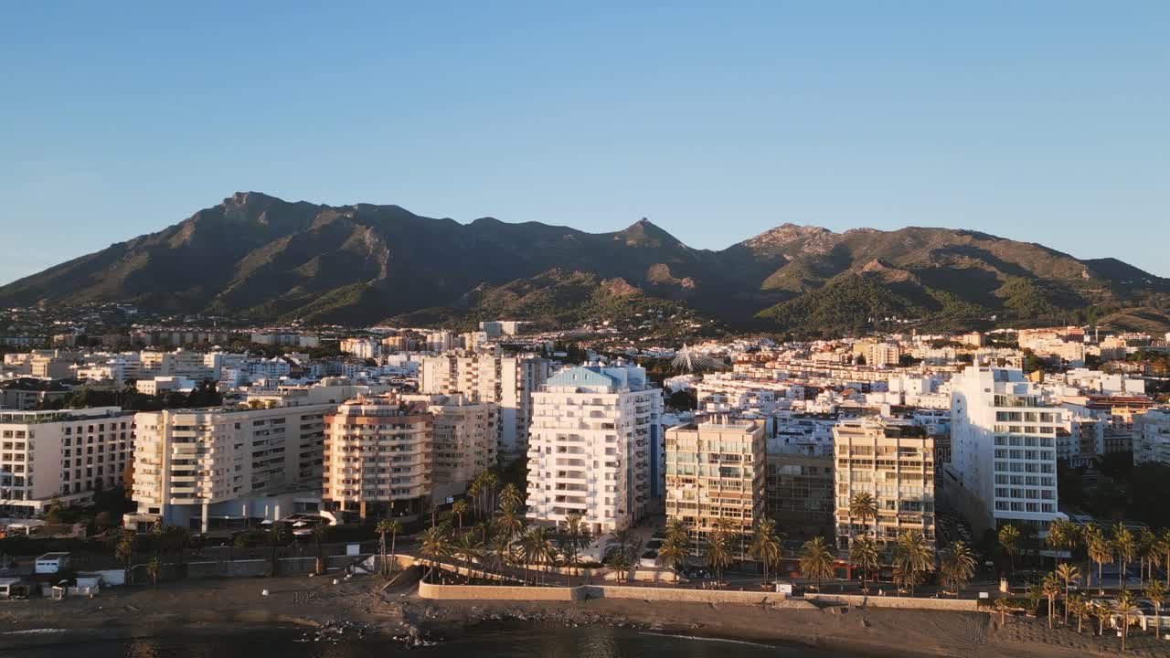 Drone che si alza verso gli edifici sul lungomare di Marbella, vista costiera con cielo azzurro limpido e le montagne della Sierra Blanca sullo sfondo, Spagna meridionale