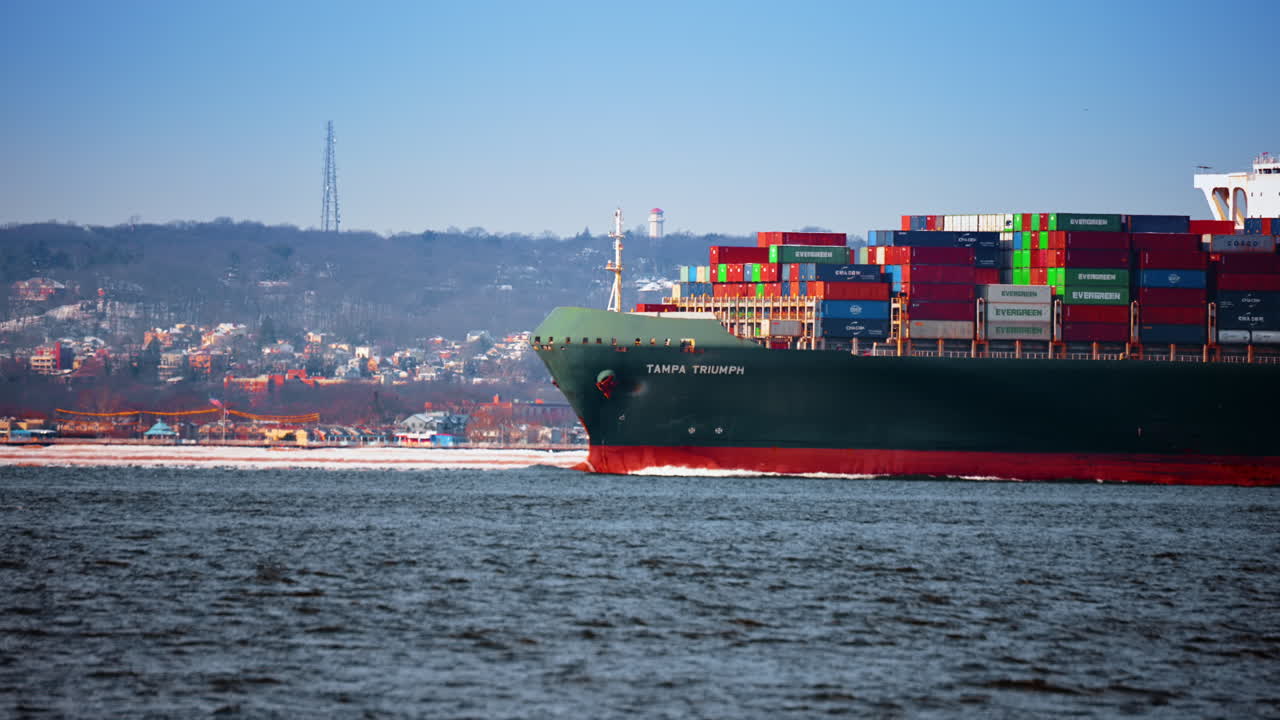 New York, USA, 25 December 2025: Container ship Tampa Triumph passing the shoreline with tower. Profile of a large green cargo vessel sailing near a hilly coast with a tall communication tower