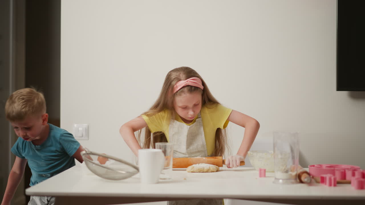 Little girl wearing ribbon on head rolling dough with wooden pin while younger boy watches closely and moves around white kitchen table, surrounded by baking tools and ingredients