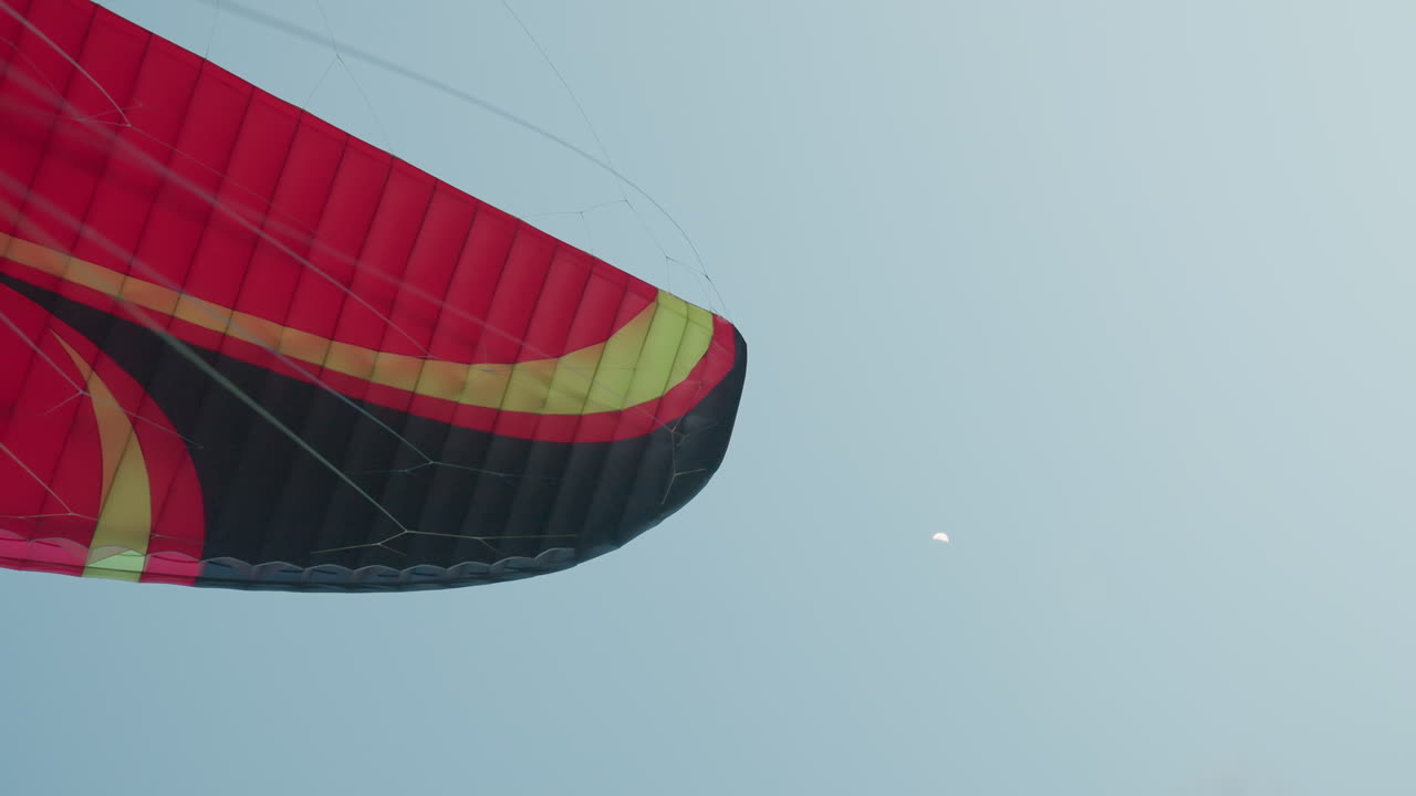 close up of red black yellow paraglider wing with visible fabric and tension lines soaring through pale blue sky with subtle moon crescent backdrop evoking serene aerial sports motion