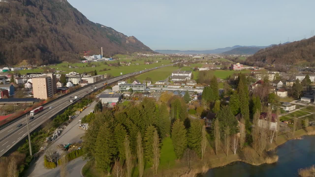 Drone flight parallel to a highway, passing over a calm pond near a small town and open green valley. Clear daylight highlights the natural scenery and infrastructure in harmony