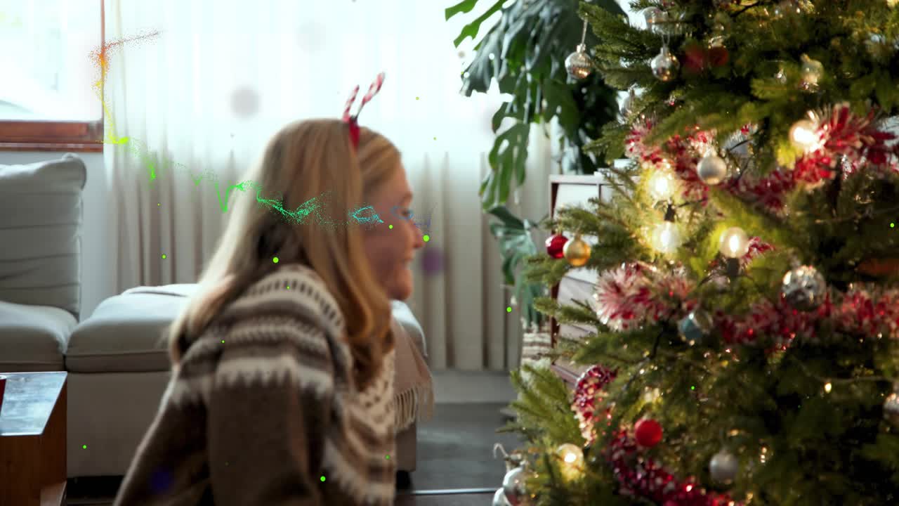 Mature woman picking card while tapping phone, donning antlers, fixing tree ornaments for holiday