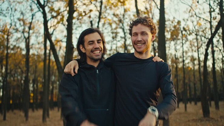 Two happy and cheerful guys posing in the autumn forest after their jog and high-fiving each other. Happy guys after a morning jog look at the camera and pose hugging in black sportswear in the autumn forest