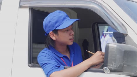 asian delivery man sitting behind wheel of car and checking customer list on clipboard before sending the package