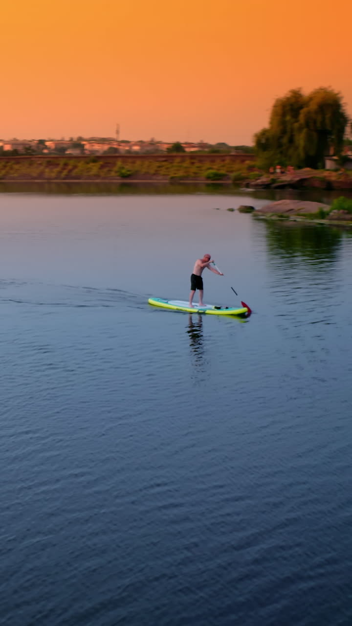 Sportsman on a paddle boat on lake at sunset. Man sailing on a board with oar along the evening river in the countryside. Extreme sport on water. Vertical video