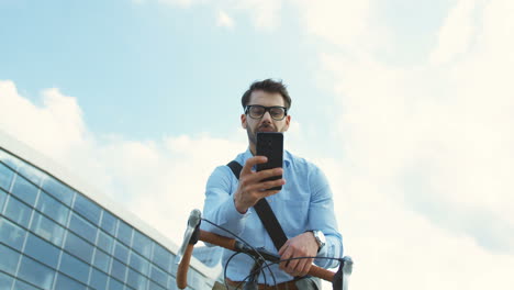 Good-looking-businessman-standing-on-the-street-and-leaning-on-his-bike,-taking-out-a-smartphone,-reading-a-message-and-smiling-cheerfully