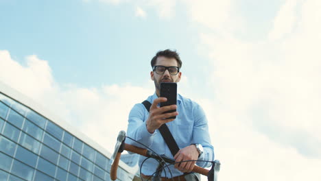 hombre de negocios apuesto parado en la calle y apoyado en su bicicleta, sacando un teléfono inteligente, leyendo un mensaje y sonriendo alegremente