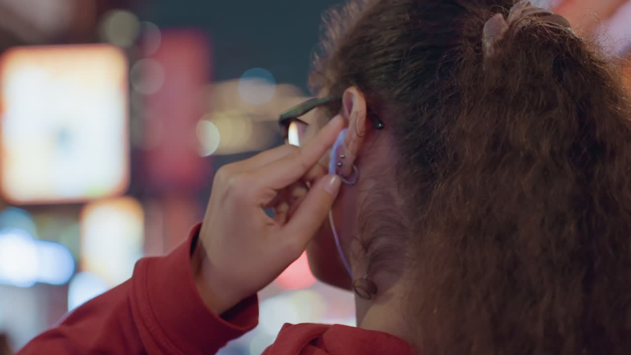Appassionato di musica afroamericano con occhiali che ascolta musica nella città illuminata al neon, atmosfera da festival serale e profilo spontaneo con auricolari, luci bokeh soffuse, sfondo di strada affollata e un'esplorazione urbana rilassata.