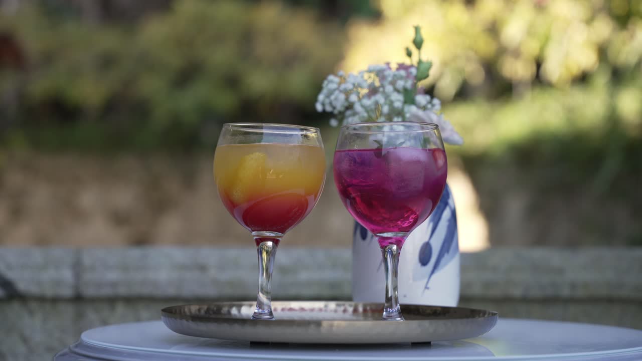 Two vibrant cocktails with fruit and ice on a tray, set on an outdoor table in soft sunlight