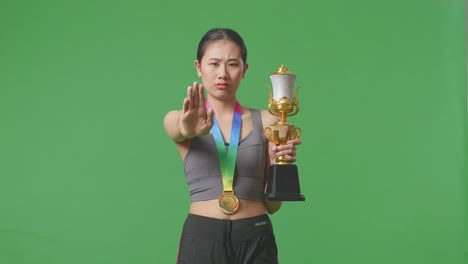 asian woman with a gold medal and trophy disapproving with no hand sign and smiling to camera on green screen background in the studio