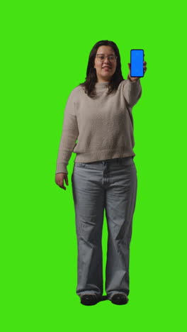 Vertical-Video-Full-Length-Shot-Of-Young-Woman-Holding-Blue-Screen-Mobile-Phone-To-Camera-Standing-Against-Green-Screen-1