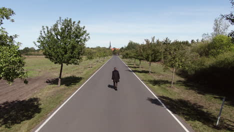 Man-walking-in-middle-of-country-road-towards-small-town,-aerial-pull-away-view