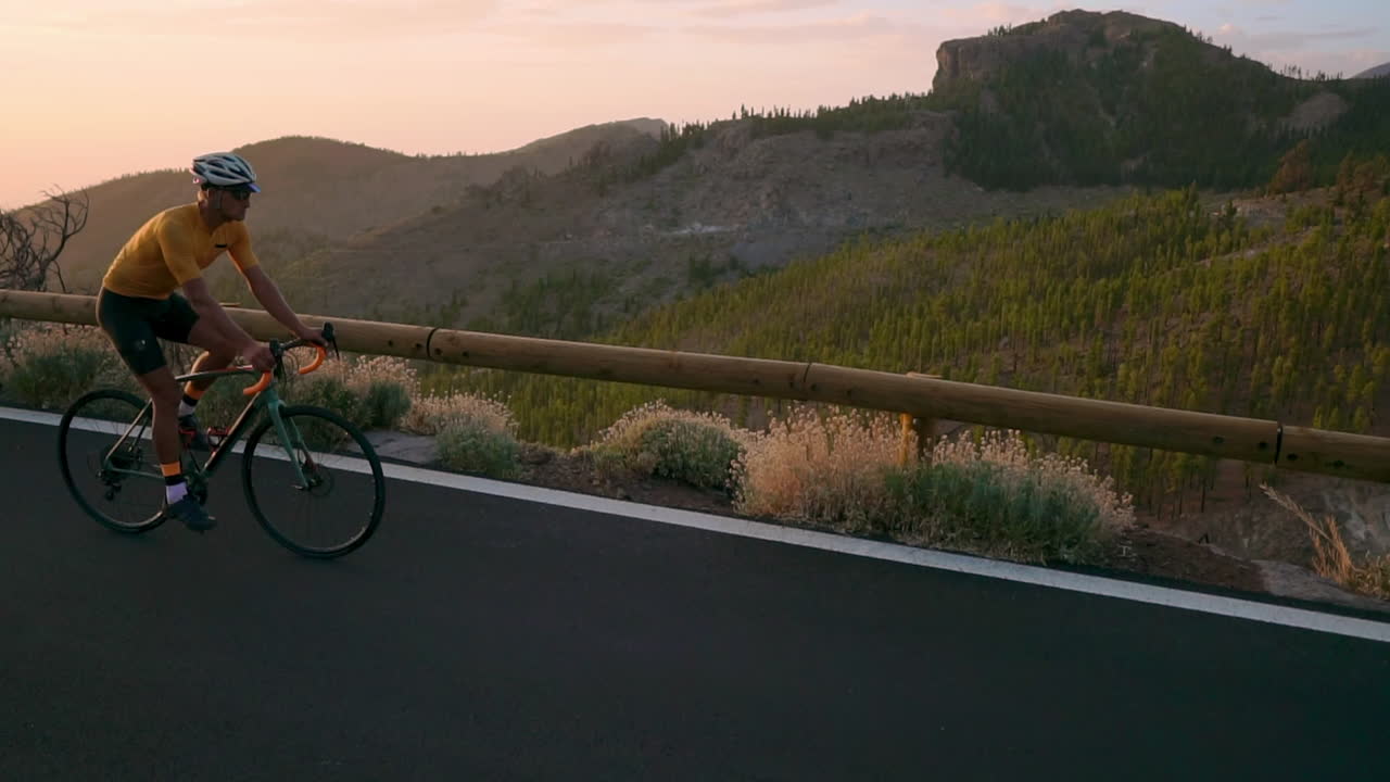 헬 ⁇ 과 스포츠 장비를 입은 전문 자전거 타는 사람이 해가 지면 화산 방향으로 산의 serpentine에 타고 있습니다. steadicam