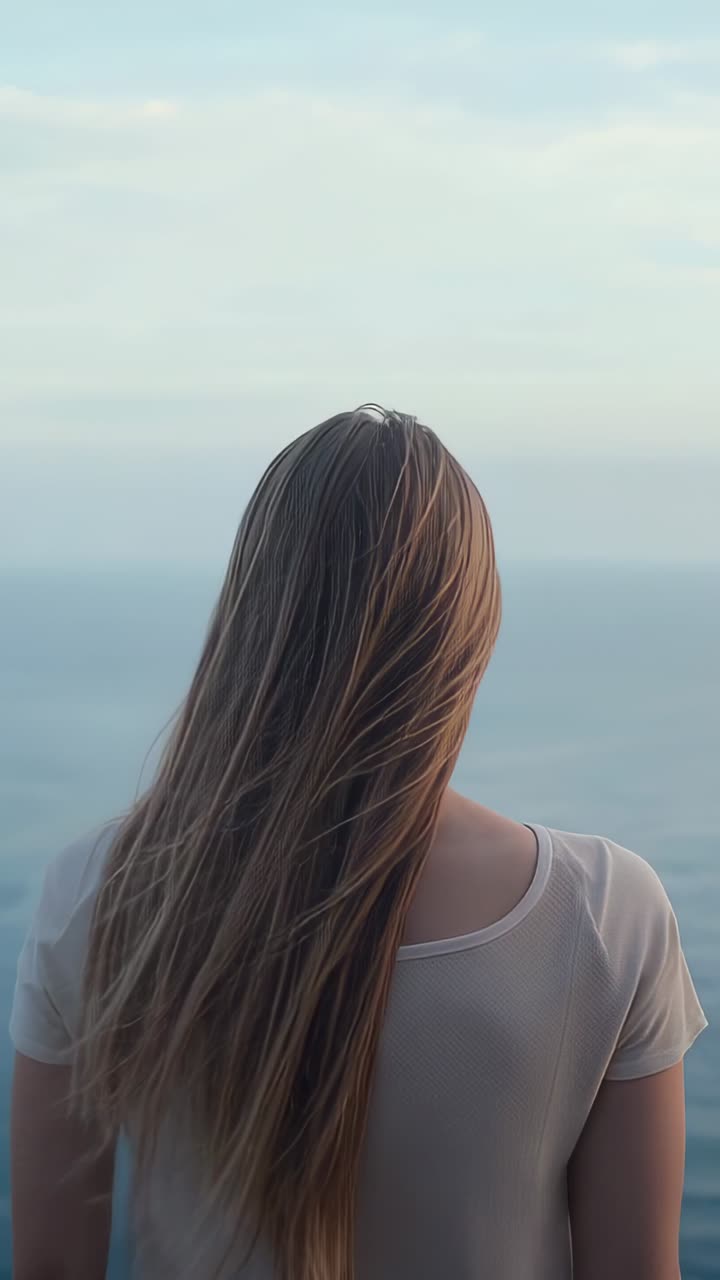 Vertical video: Gazing woman feeling breeze on coastal cliff seeking calm, hair flowing to horizon