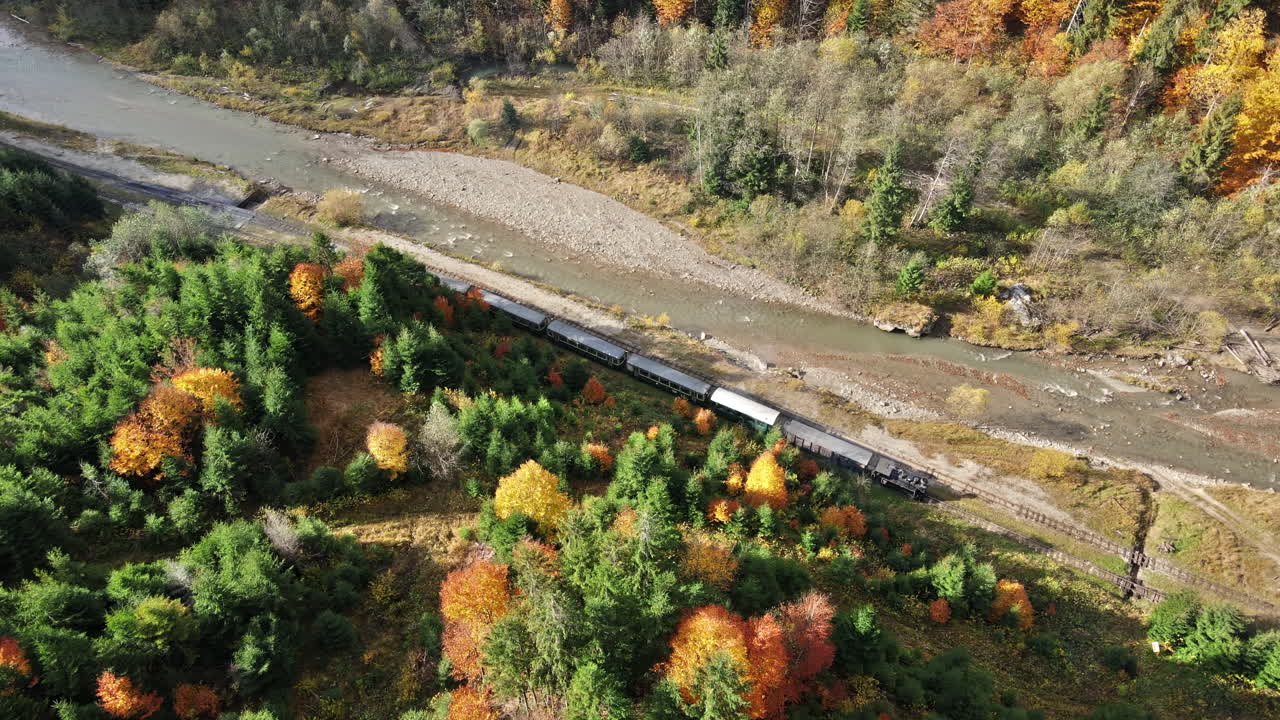 Aerial drone view of the moving steam train Mocanita in a valley along a river, hills covered with yellowing forest, Romania