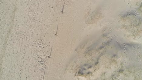 Long-fence-post-shadows-stretch-out-across-a-smooth-sandy-beach-covered-in-footprints,-aerial-god-view