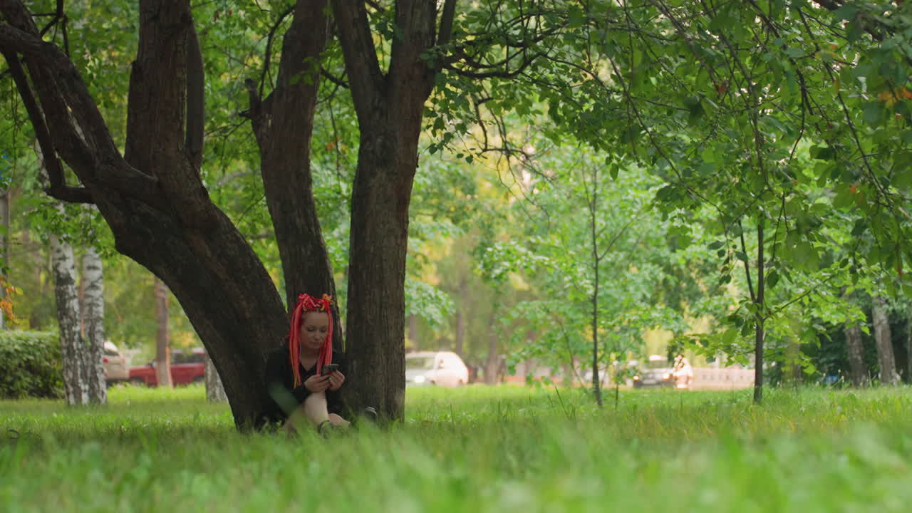 잎이 무성한 공원에서 땋은 머리를 한 십 대가 나무 줄기에 기대어 있고, 낮은 각도의 풀밭이 전경에 보이며, 빨간색 백팩이 보이고, 사색에 잠긴 표정, 조용한 도시의 녹지, 그리고 친밀한 인물 사진 구도
