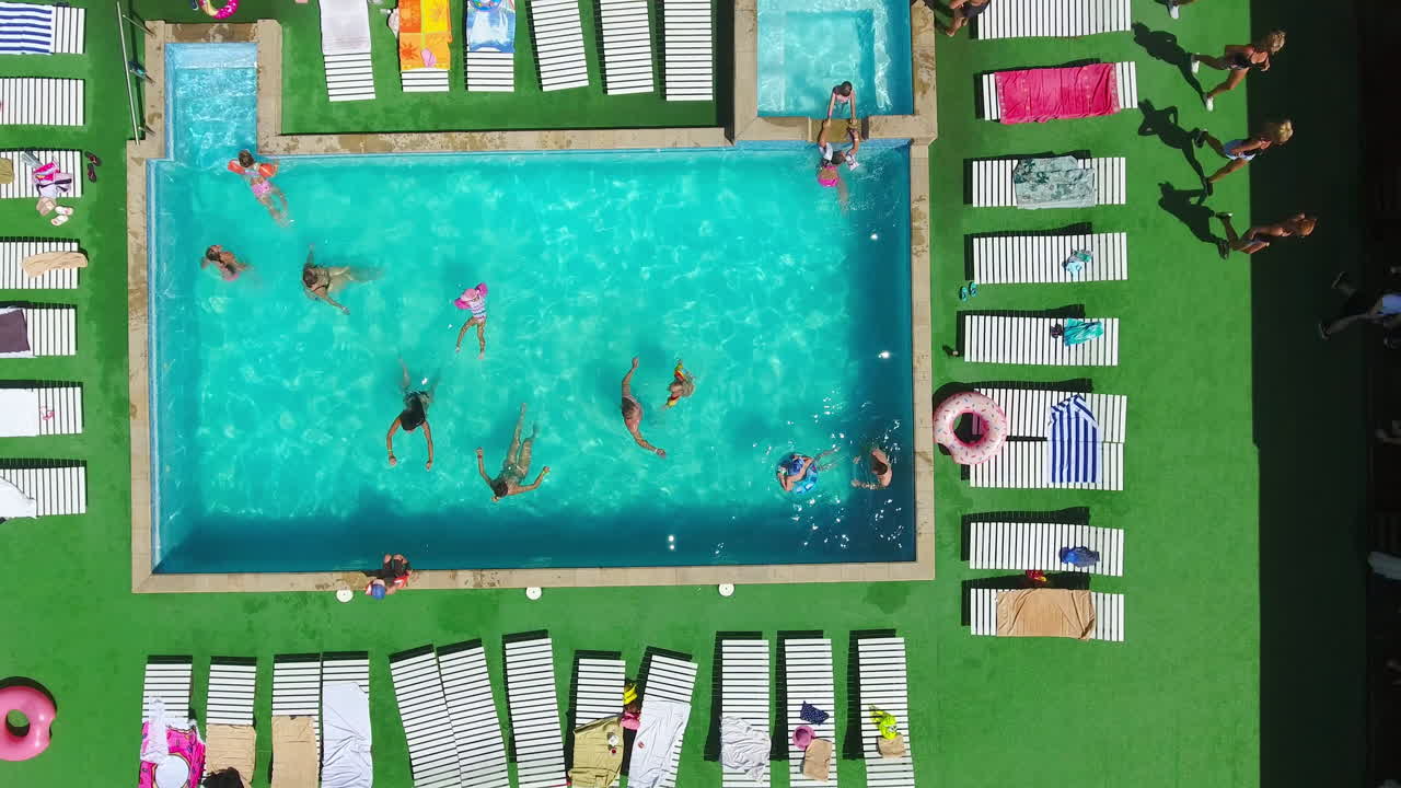 Happy people swimming in the blue pool, doing aerobics around and sunbathing on the chaise-longues. Drone footage going up over the spa resort territory.