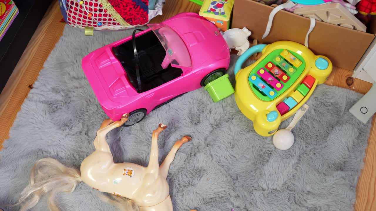 Draufsicht auf eine unordentliche Spielecke für Kinder mit einem Spielzeugpferd, einem knallrosa Auto und einer Babylaufhilfe, die auf einem flauschigen grauen Teppich neben Holzregalen und -kisten verstreut sind