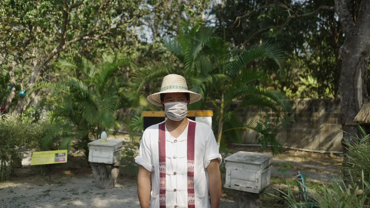 A man wearing a straw hat and face mask stands in front of beehives at an apiary