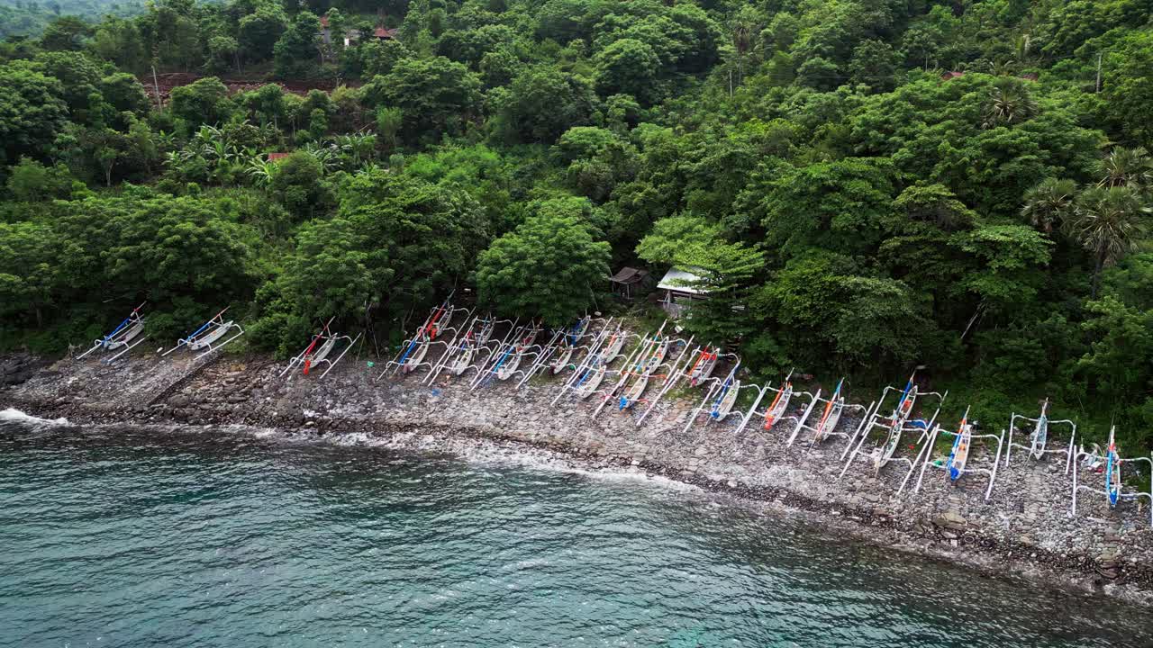 Veduta aerea con drone di colorate barche da pesca allineate lungo una spiaggia rocciosa a Bali, incorniciate da fitta vegetazione della giungla e dolci colline costiere accanto a calme acque oceaniche.