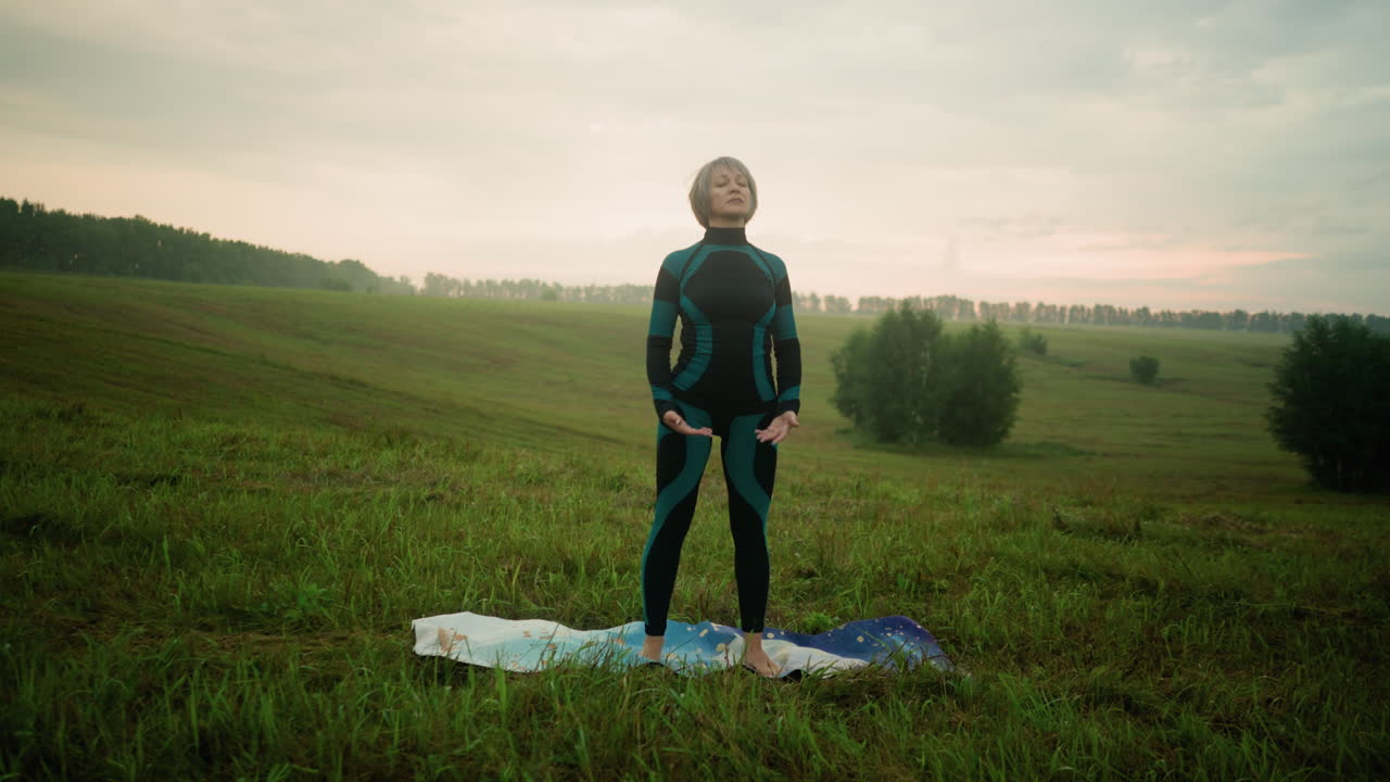 женщина среднего возраста в зелено-черном костюме и леггинсах стоит на коврике в травяном поле, готовясь к йоге под спокойным небом на закате, в окружении деревьев в открытой сельской местности