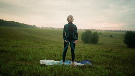 mujer de mediana edad en traje verde y negro y leggings de pie en una alfombra en el campo cubierto de hierba preparándose para el yoga bajo el cielo sereno al atardecer, rodeada de árboles en el campo abierto