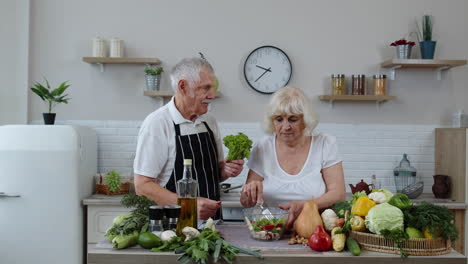 Ältere-Großeltern-In-Der-Küche.-Lustiger-Opa-Macht-Witze-über-Oma.-Legte-Einen-Salat-Um-Ihren-Kopf