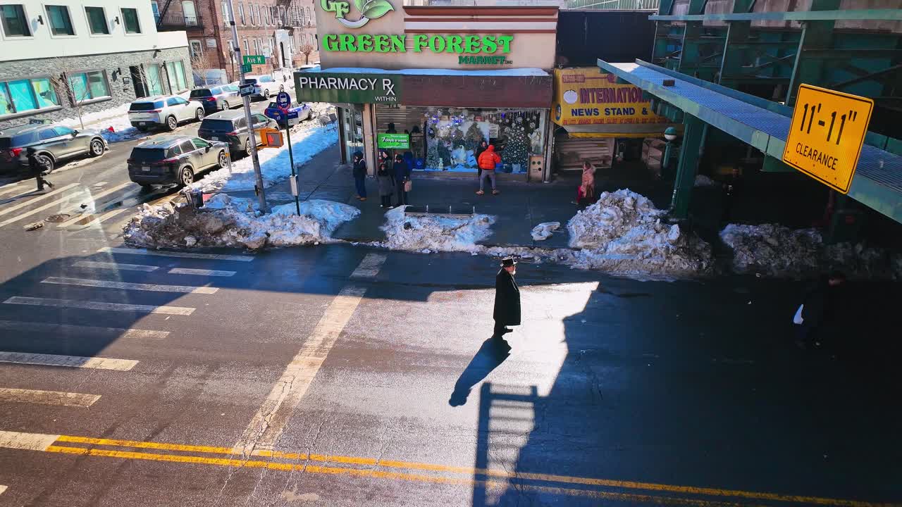 New York, USA, 25. Dezember 2025: Orthodoxer jüdischer Mann überquert eine städtische Straße in New York. Vogelperspektive eines Fußgängers in traditioneller schwarzer Kleidung, der eine verschneite Stadtkreuzung nahe einem Markt überquert.