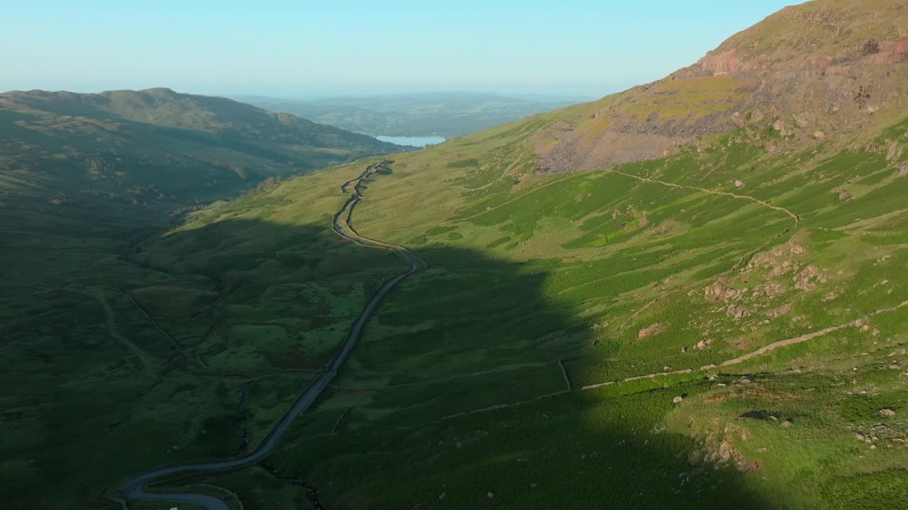 Sunrise light on mountainside with mountain pass road heading towards distant lake Windermere. Summer. Kirkstone Pass, Lake District, Cumbria, UK