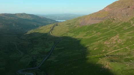 sunrise light on mountainside with mountain pass road heading towards distant lake windermere. summer. kirkstone pass, lake district, cumbria, uk