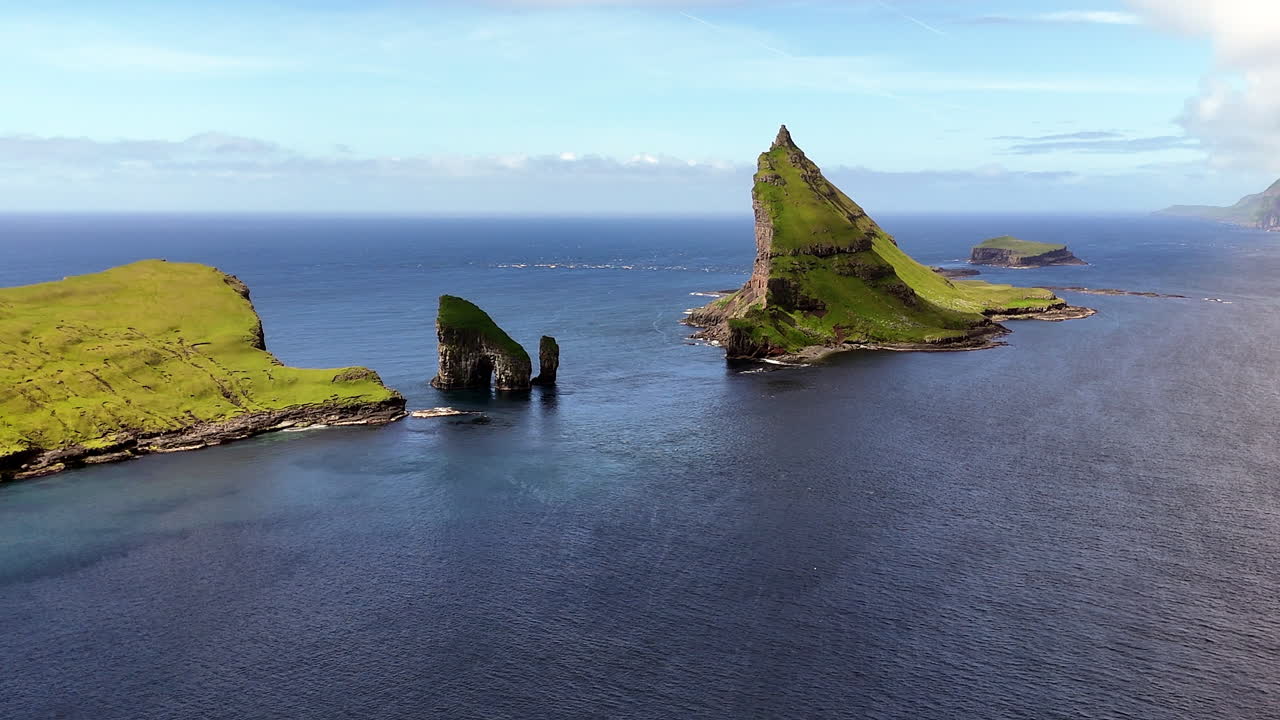 Cinematic aerial view of Drangarnir sea stacks rising dramatically from the Atlantic Ocean near Vágar, Faroe Islands, showcasing rugged cliffs, lush green slopes, and misty Nordic seascape