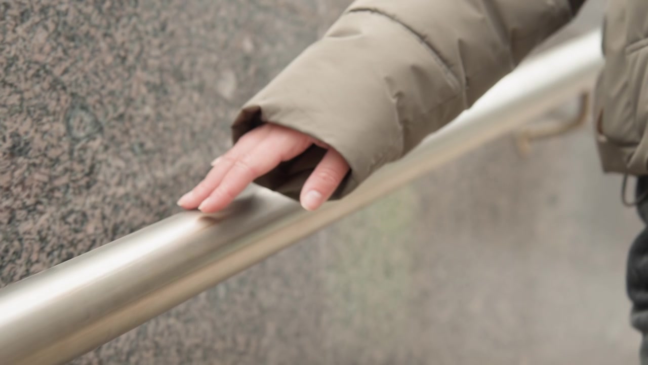 Hand resting on metal handrail by stair entrance, closeup of bare fingers and puffer jacket sleeve, commuter pausing for balance on chilly day, polished steel rail and urban transit vibe