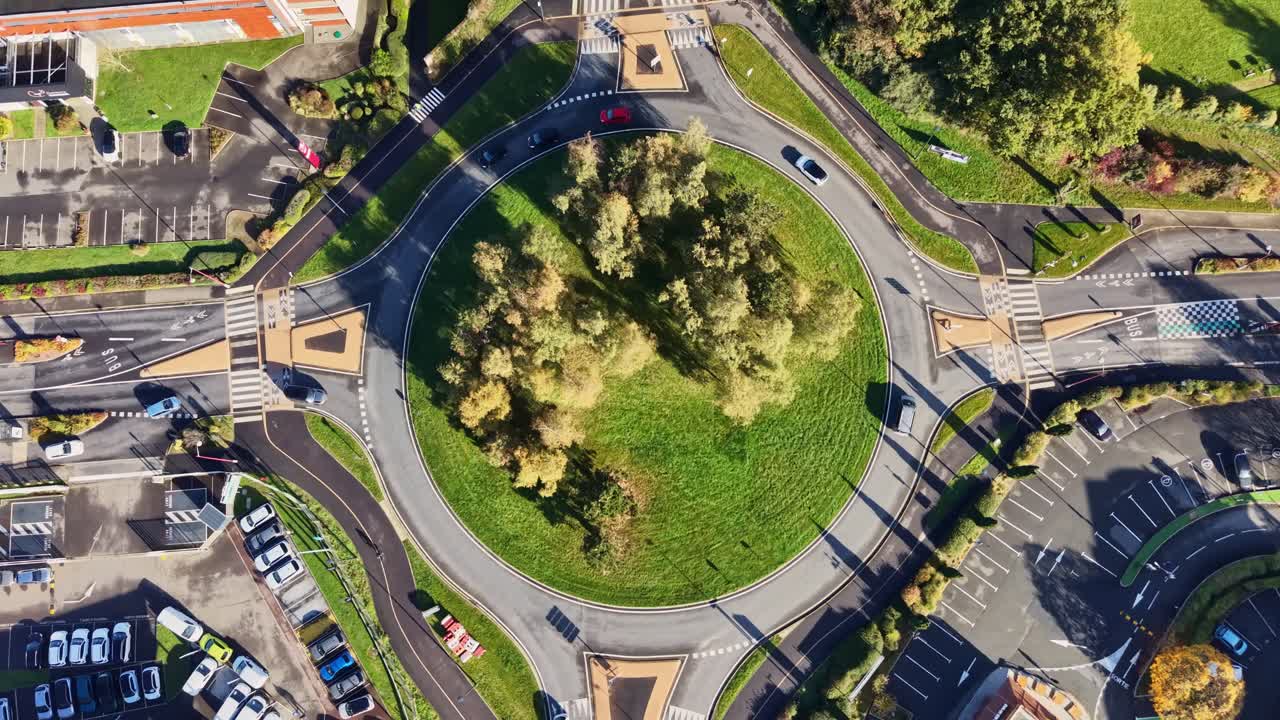 Drone shot flying slightly forward with a top-down view of a roundabout in Cesson-Sévigné, France, showing cars, trees, surrounding buildings, and parking lots