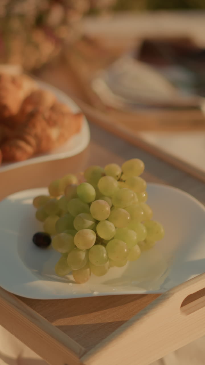 primer plano de la configuración de picnic en una mesa de madera con platos de uvas verdes frescas, croissants y bocadillos bajo la luz solar natural, los platos están artísticamente dispuestos creando un aire libre relajado